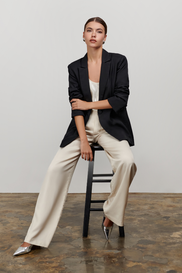 Woman sitting on a stool wearing a black blazer and beige pants against a plain background